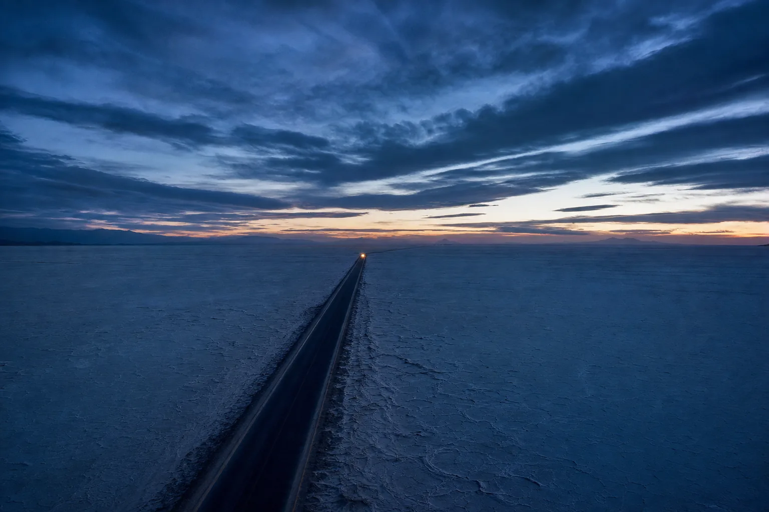 An empty road crossing a vast salt flat at dusk with a single distant headlight